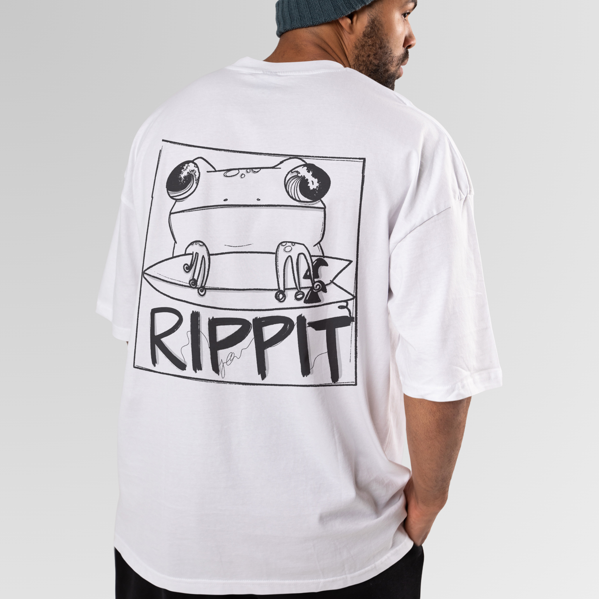 back view of a model wearing a cool surf shirt featuring a design of a frog holding a surfboard over text that says 'rippit'