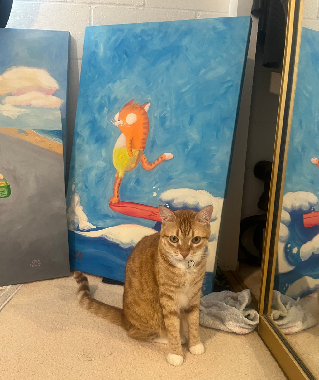 An orange tabby cat wearing a small bell sits on a beige carpet in front of a large vertical canvas painting. The painting depicts an orange cat surfing on white waves under a blue sky.
