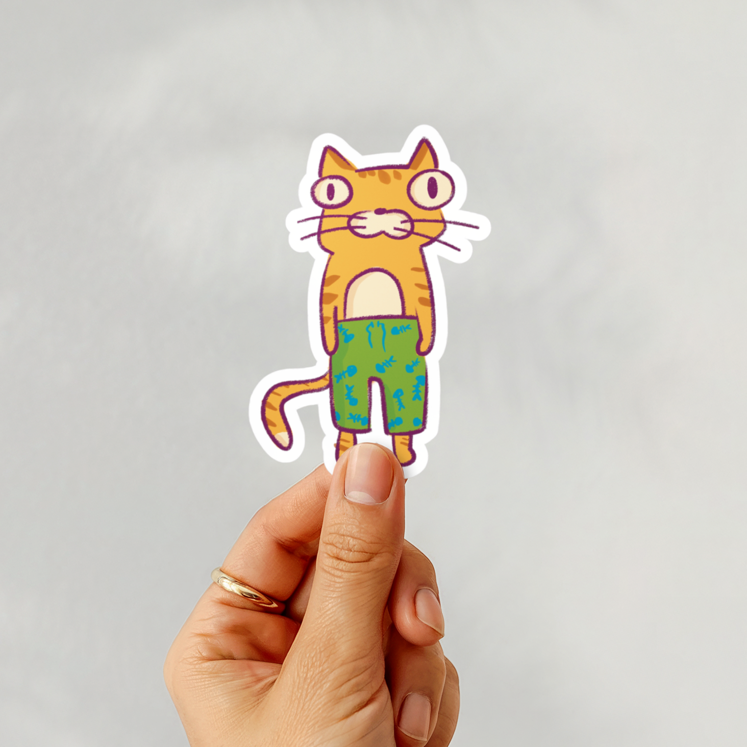 Hand holding a sticker of a cat wearing green pants against a gray background