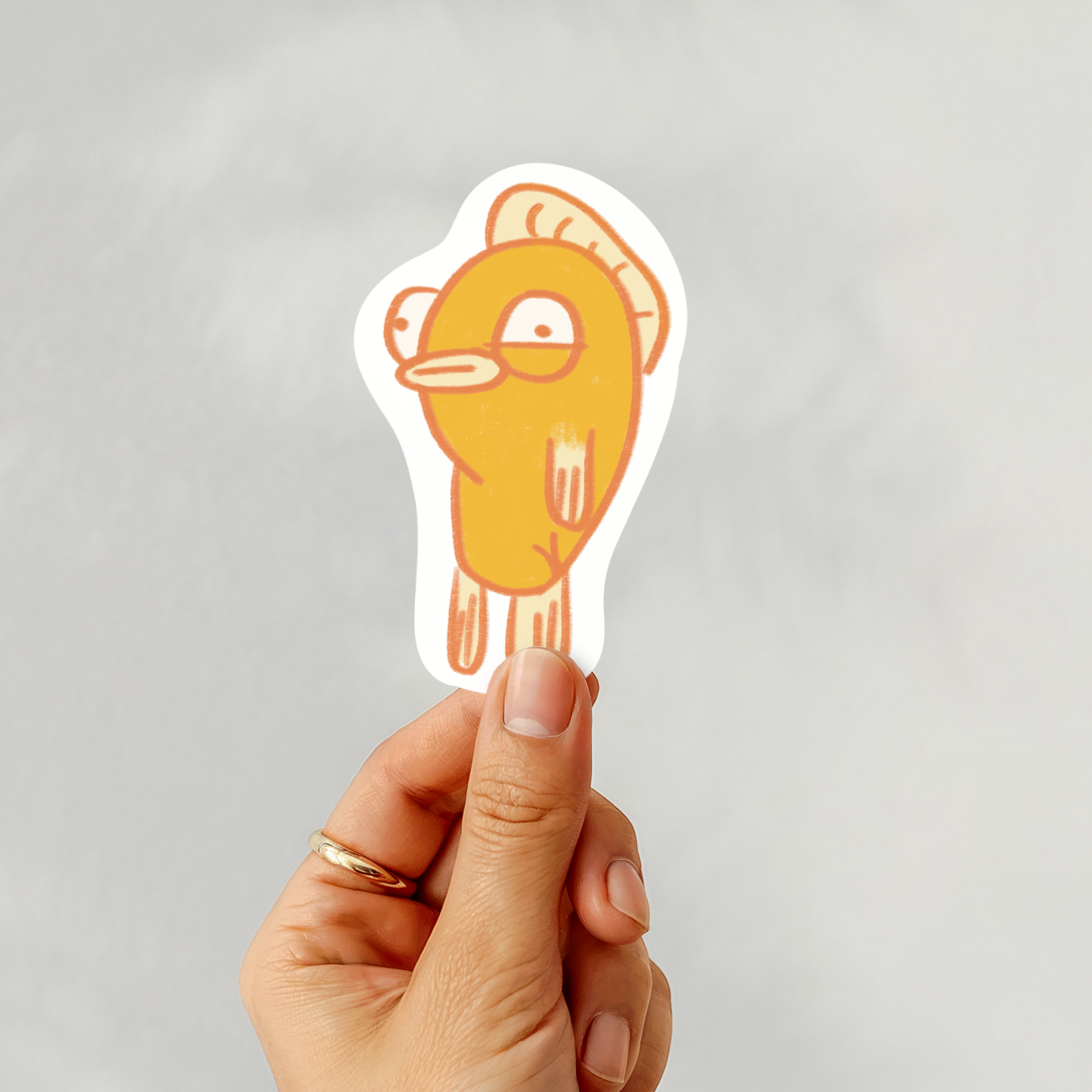 Hand holding a yellow cartoon-like fish character sticker against a gray background