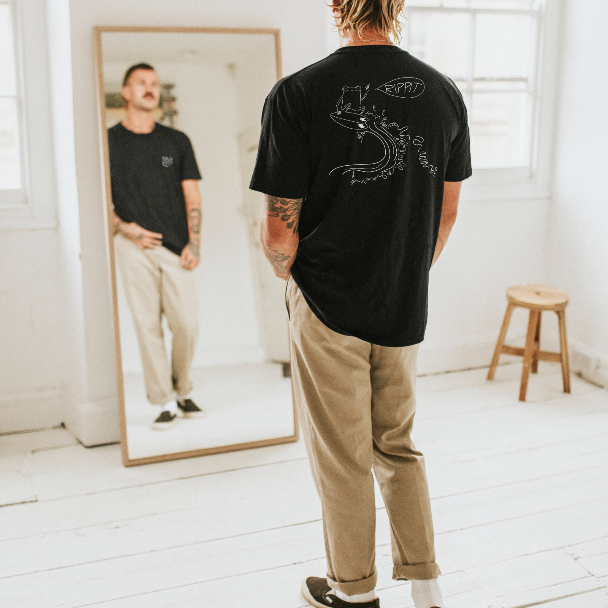 male surfer model wearing a black surfjoy t-shirt featuring a design of a frog catching an air on a wave with a speech bubble that says 'rippit'
