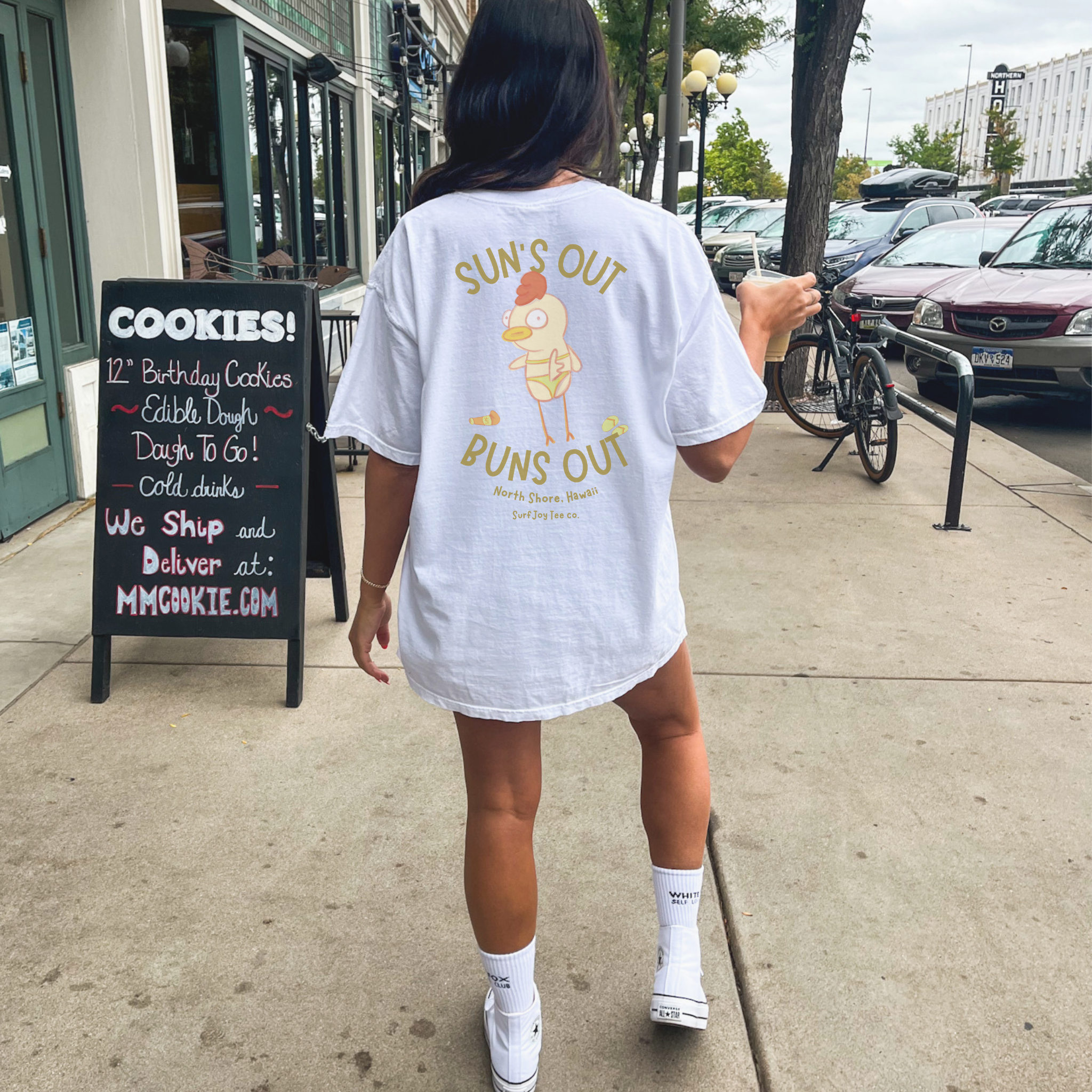woman wearing a funny beach t-shirt with a chicken in a bikini by text that reads 'suns out buns out' while holding an iced coffee 