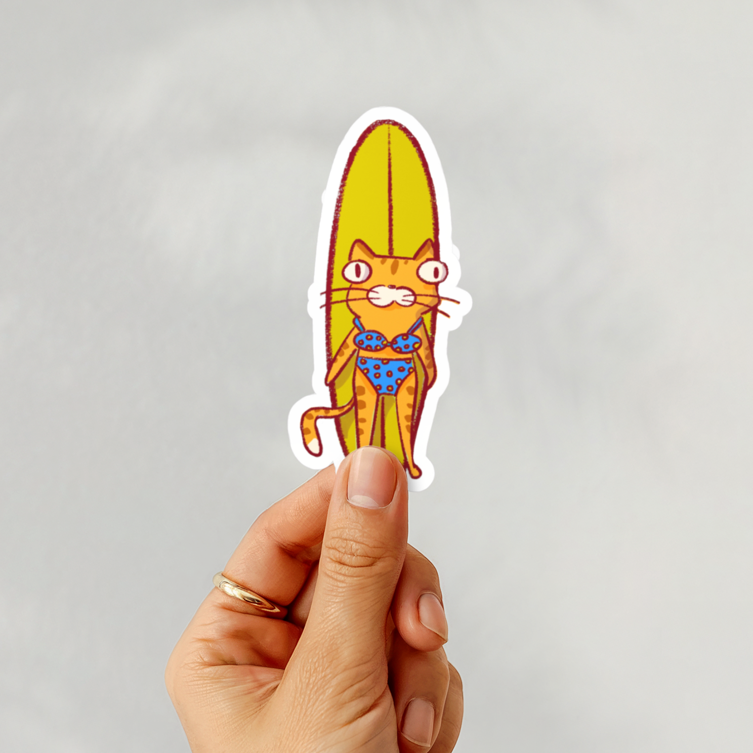Hand holding a colorful sticker of a cat on a surfboard against a gray background