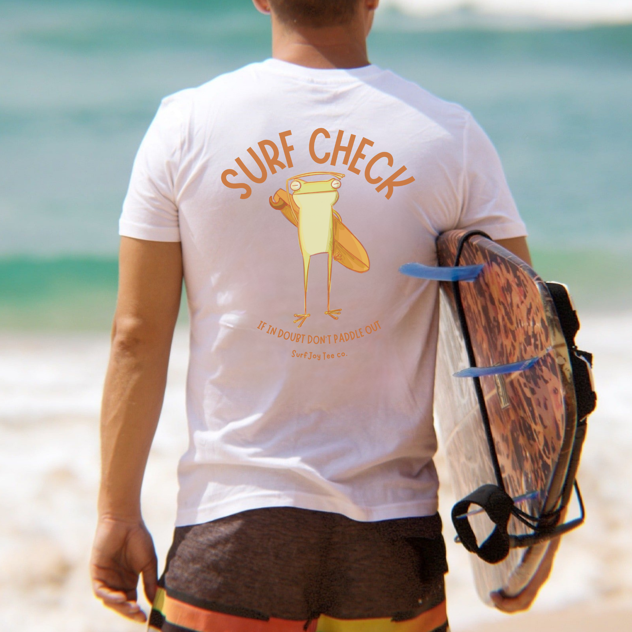 man in board shorts holding a surfboard wearing a surfjoy t-shirt featuring a funny design of a frog checking the surf conditions next to text that says 'if in doubt, don't paddle out'