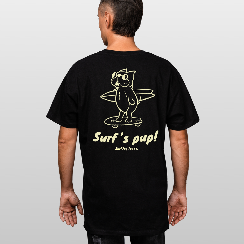 back view of a male model in a black t-shirt with a design of a dog skating to the beach carrying a surfboard over text that reads 'surf's pup!'