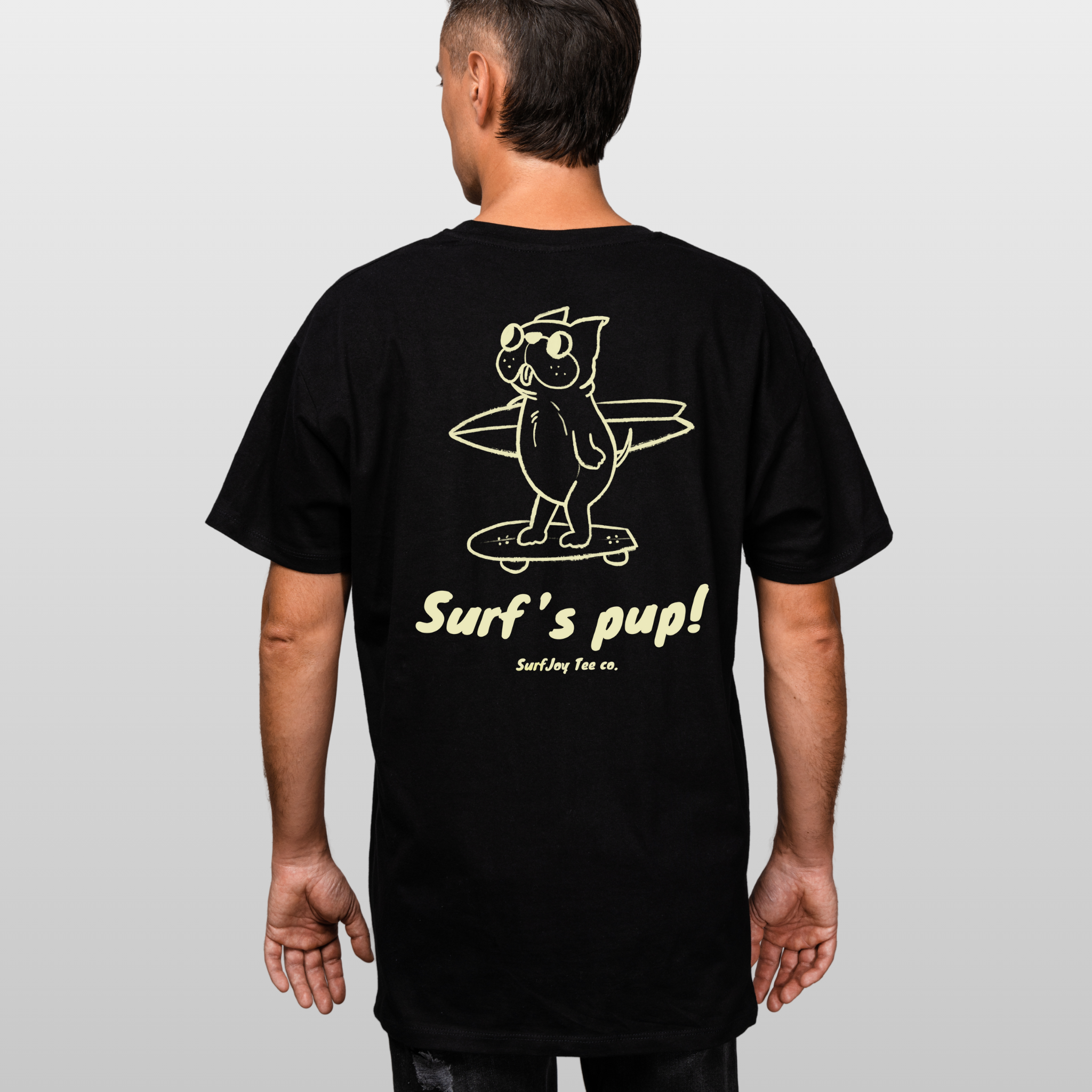 back view of a male model in a black t-shirt with a design of a dog skating to the beach carrying a surfboard over text that reads 'surf's pup!'