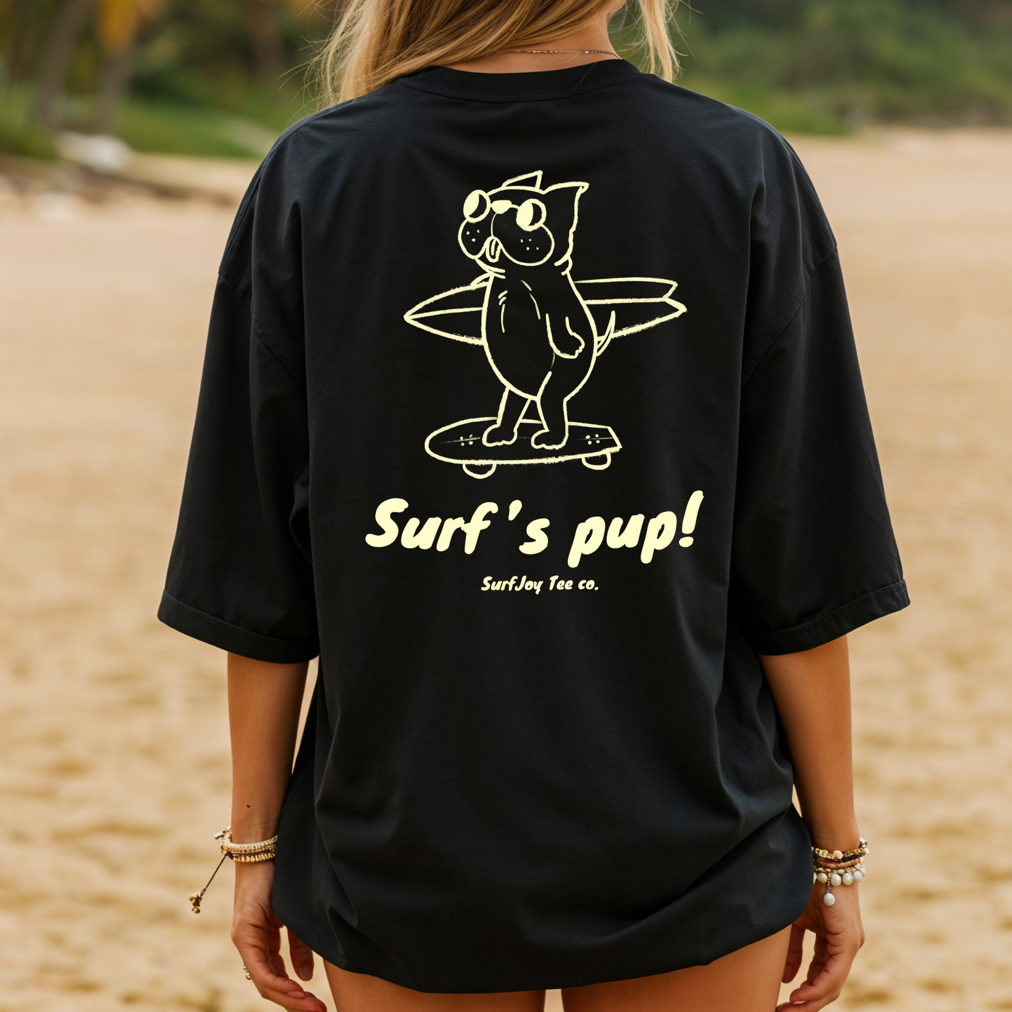 back view of a woman wearing a black t-shirt with a design of a dog skating to the beach carrying a surfboard over text that reads 'surf's pup!' on the beach