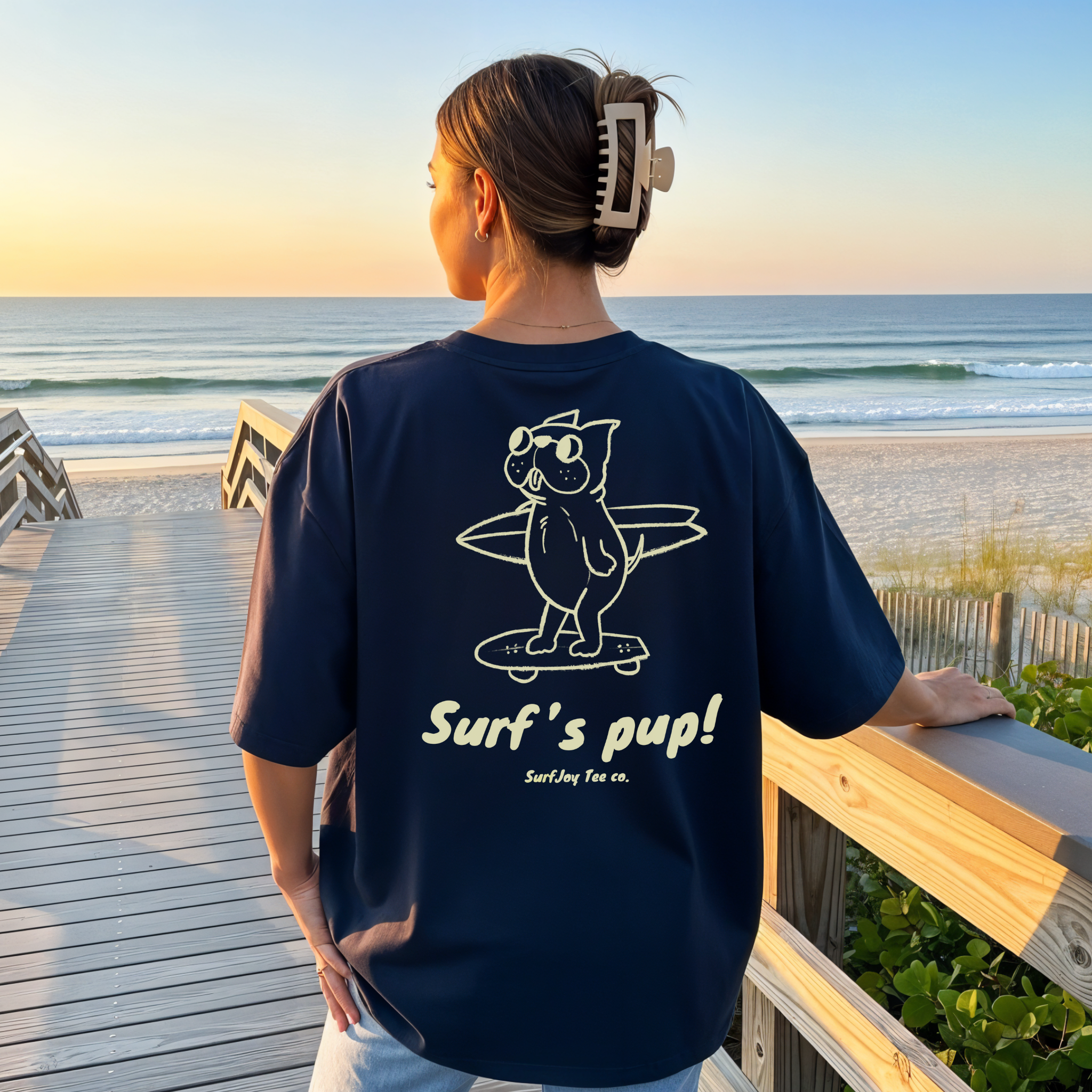 woman on a beach wearing a navy blue surfer t-shirt with a design of a dog skating to the beach carrying a surfboard over text that reads 'surf's pup!'