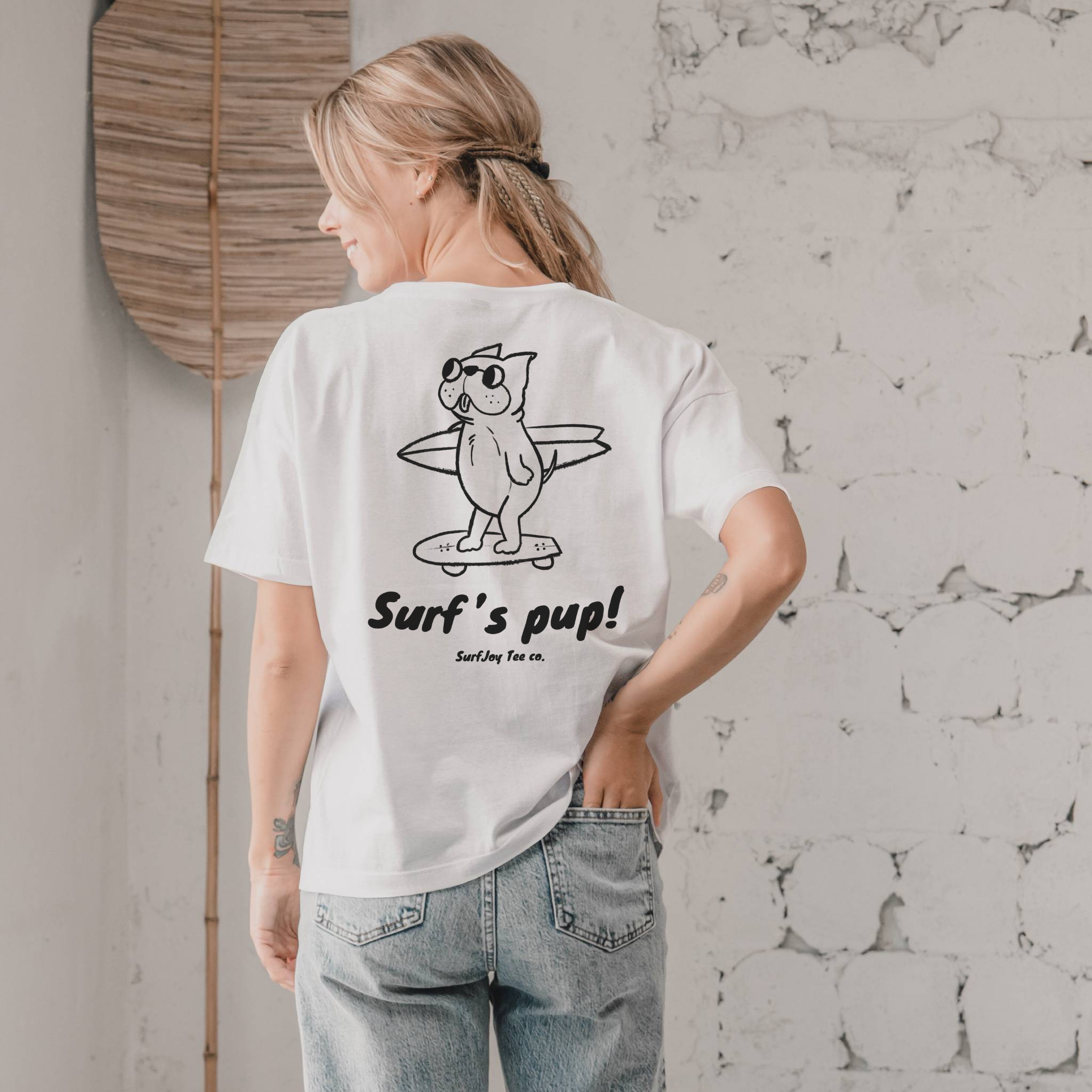 back view of a woman wearing a white surfer t-shirt with a design of a dog skating to the beach carrying a surfboard over text that reads 'surf's pup!'