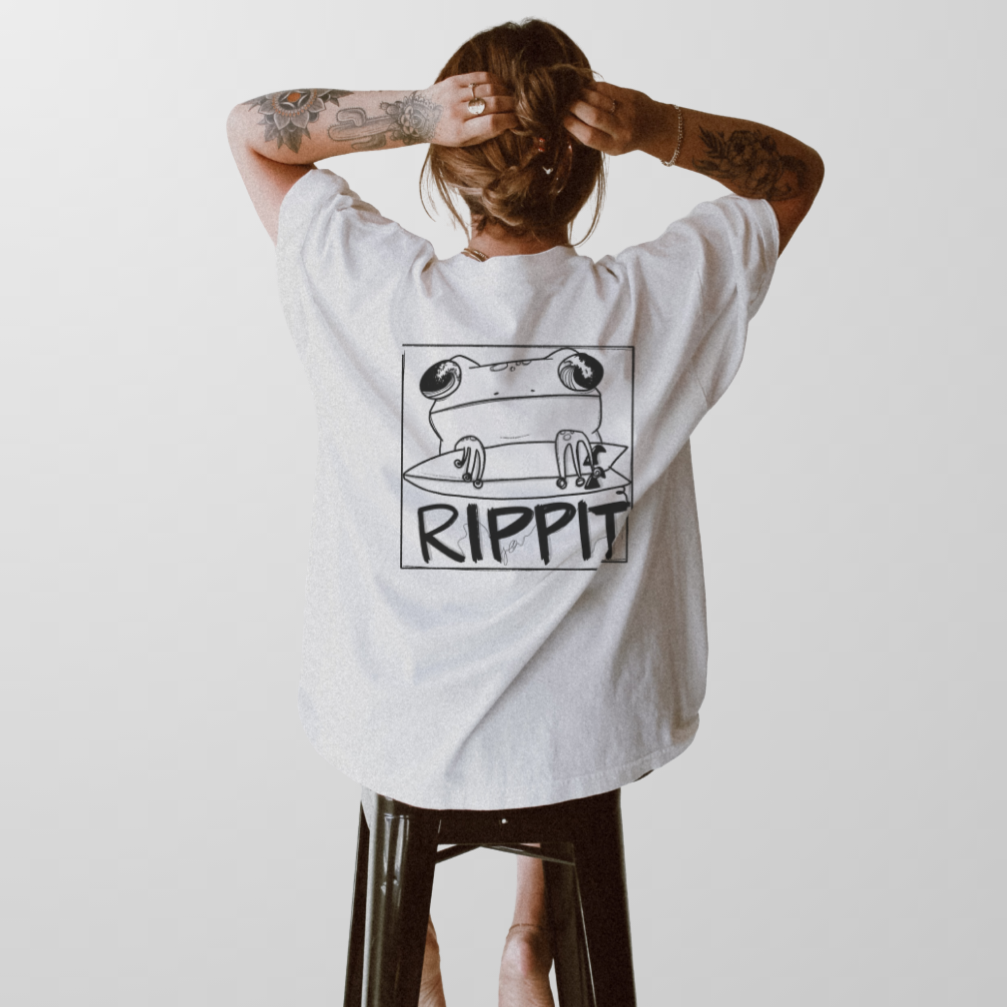 woman wearing an oversized surfjoy t-shirt is seated on a stool showing the back printed design of a frog holding a surfboard over text that says 'rippit'