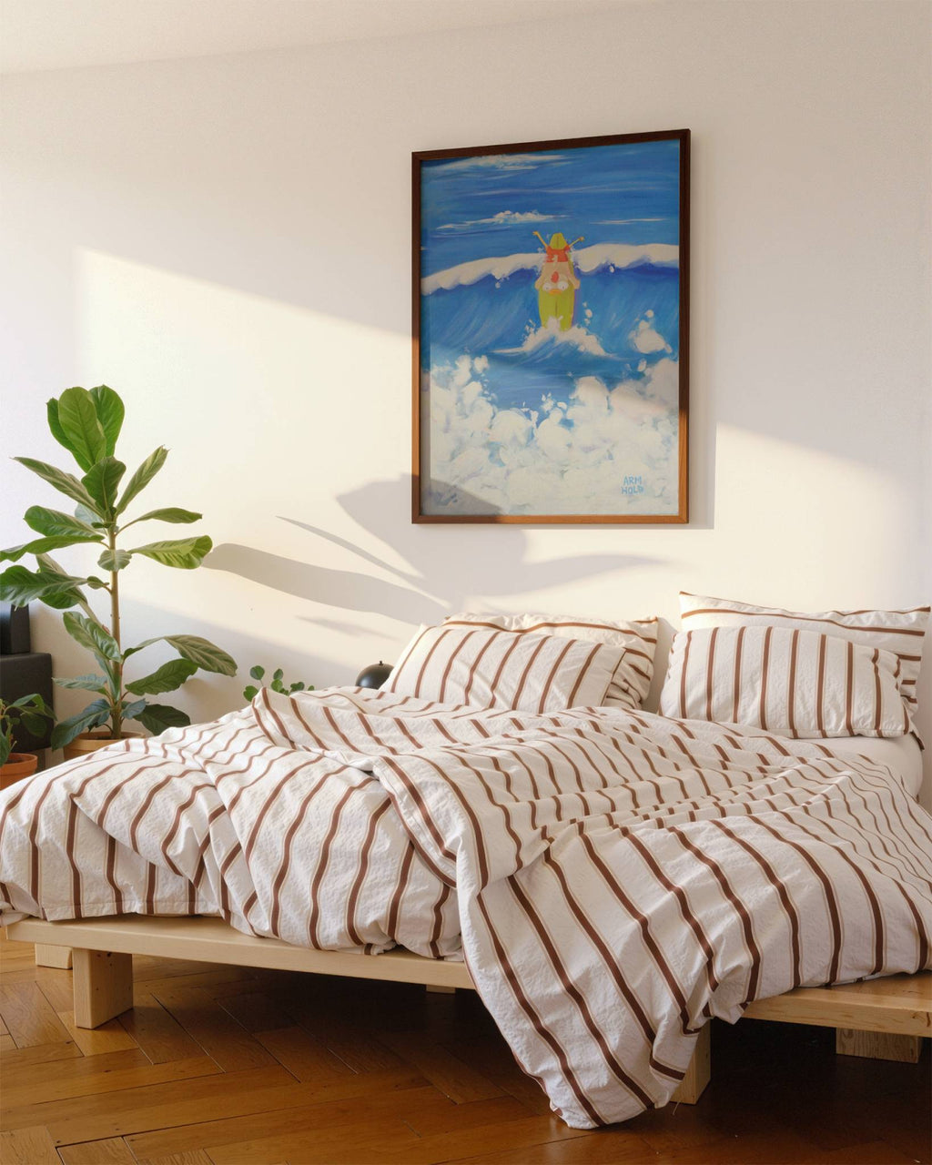 A styled bedroom scene with a bed dressed in cream bedding featuring brown vertical stripes, set against a white wall with a framed painting of a surfer. A green potted plant is on the left, and warm sunlight casts shadows across the wall and bedding.